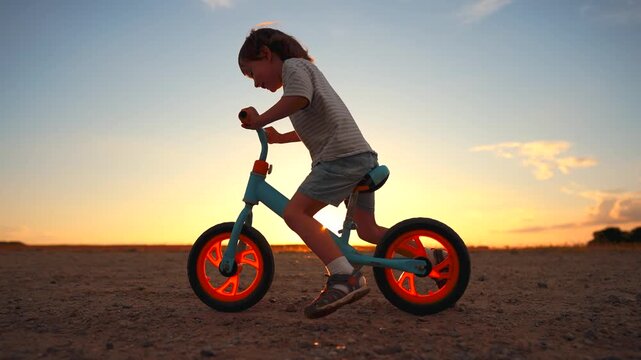 A girl rides a balance bike against a sunset backdrop. The girl is focused on balancing as the colors of the sunset create a warm atmosphere for her ride.