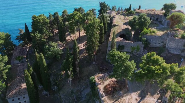 Cinematic high angle drone view of a medieval stone fortress with historic ruins, green pine trees, and old buildings on a coastal hill side.