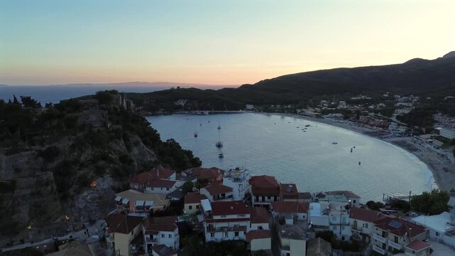 Cinematic aerial view of a scenic coastal town revealing a large sandy beach bay with anchored boats during twilight.