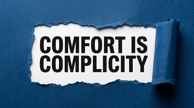 The phrase Comfort Is Complicity written on white paper behind a torn blue surface.