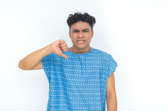 Young male patient in a hospital gown gives a thumbs down gesture with an angry expression, expressing dissatisfaction, frustration, or negative feedback.