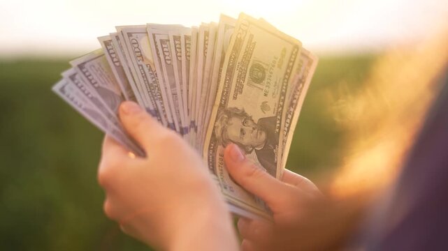 A woman holds a fan of money, featuring various denominations, in her hands. The sunlight illuminates the cash, creating a bright scene with a green background.