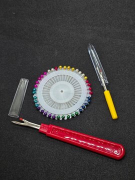 Round wheel of colorful pearl headed pins and two seam rippers against textured black background, concept of professional tailoring and needlework