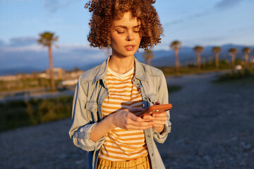 Fototapeta premium A woman in a denim jacket and striped shirt stands outdoors at golden hour, focused on her smartphone. A breezy coastal backdrop highlights casual, stylish everyday life.