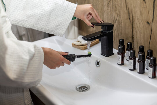 Hand rinsing toothbrush under bathroom faucet. Person in a white robe rinses an electric toothbrush under running water from a black bathroom faucet beside small bottles and sink accessories.
