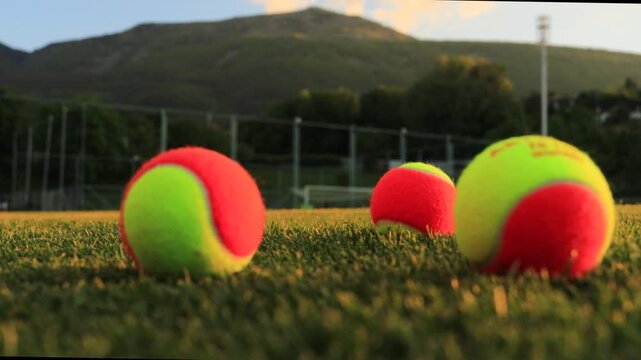 A womans hand throws orange tennis balls onto the green grass of a stadium at sunset, with beautiful mountains and a sports stadium in the background.