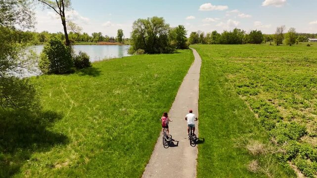 Couple cycles paved path through nature with water views, St Lawrence Parks, Ontario. Active lifestyle, healthy living, outdoor recreation, Canadian landscape.