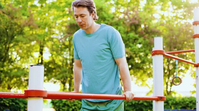 Active man doing calisthenics dips on parallel bars. Outdoor fitness workout and strength training exercise in a sunny summer park. High quality 4k footage