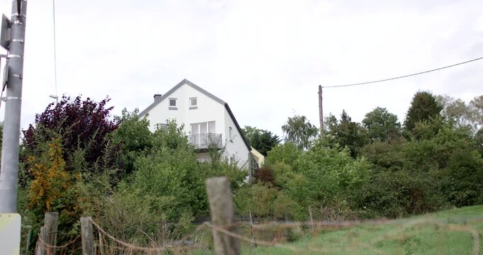 Distant view of a white two-story house surrounded by green trees and a fence under an overcast sky
