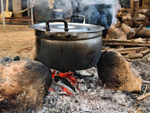 A scene of traditional Thai rural cooking featuring a steaming metal pot on a primitive firewood stove made of stones. Showing smoke, ashes, and a local person in the background, representing authenti