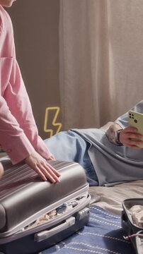 Vertical video of young adult Caucasian couple struggling to close overfilled suitcase on bed. Footage is perfect for travel agencies, luggage brands, and vacation planning campaigns