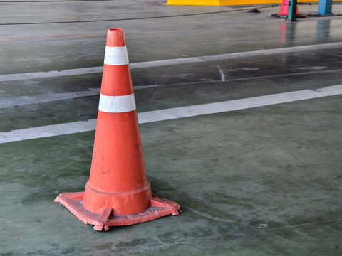 An orange plastic safety cone with white stripes placed on a grey concrete floor inside a building or parking area. Concept for caution, workplace safety, traffic control, and warning.