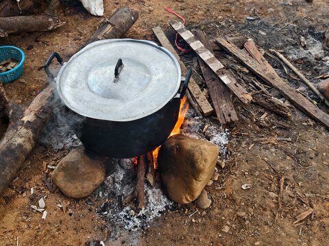 A high-angle shot of a large aluminium cooking pot boiling over an open wood fire supported by stones. Surrounded by scattered firewood, representing outdoor camping and rural cooking methods.