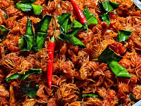 A close-up background of golden crispy deep-fried shrimp heads or chins. Garnished with fried kaffir lime leaves and red chilies, a popular Thai snack, appetizer, or street food.