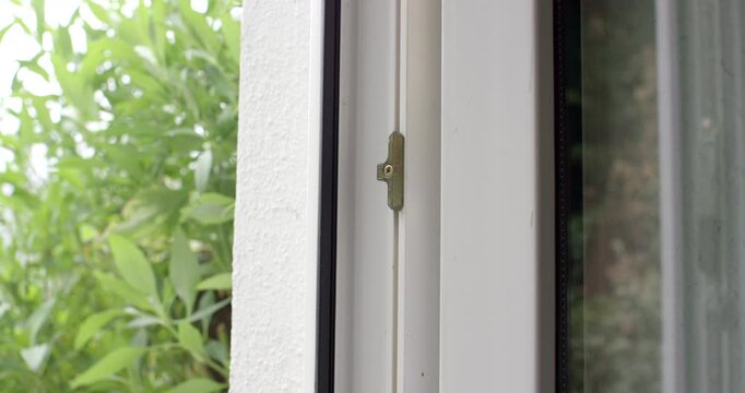 Close-up shot of a white window frame with a metal lock element and green bushes outside