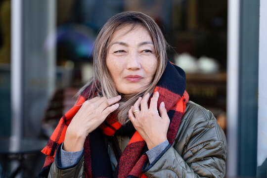 Woman stands outside a cafe in winter wearing a scarf and looking thoughtful while the wind blows