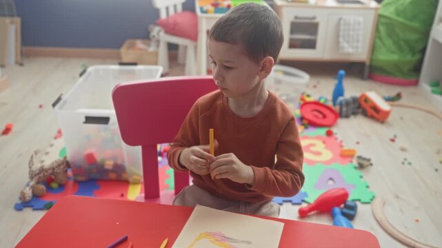 Boy drawing with yellow crayon, holds pencil in right hand and rests left hand on paper at red table in building playroom scattered with foam numbers and toys; concentration childhood.
