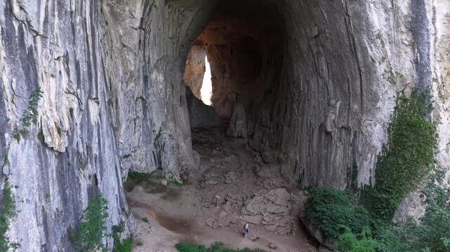 A stunning aerial or drone shot of a massive, open limestone cave. The camera slowly moves toward the cavern's immense entrance, revealing natural rock formations, majestic vertical cliffs,