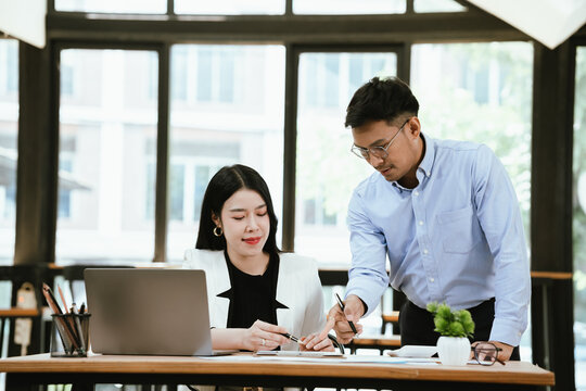 Smiling business colleagues collaborating at a modern office, reviewing work on a laptop while sharing ideas over coffee, highlighting teamwork, productivity, and professional communication.