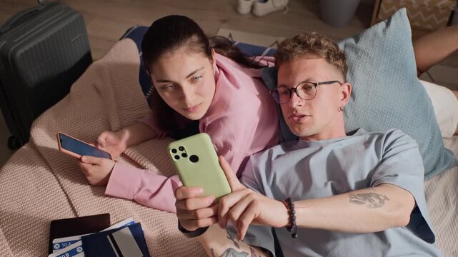 View from above of Caucasian young couple lying on bed, using smartphones, planning vacation together. Concept video for travel booking apps, mobile reservation services, and digital tourism marketing