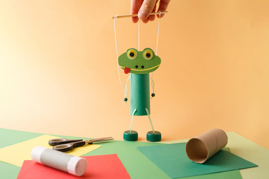 Hand holding frog marionette made from cardboard tube, paper, strings and bottle caps with scissors, glue stick, colored paper sheets and empty cardboard roll. DIY recycling craft puppet making