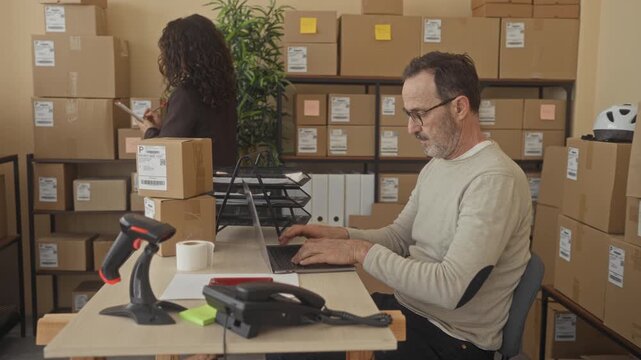 Man typing on laptop while woman gestures with clipboard beside stacked parcels, barcode scanner and packing tape in a building shipping area; productive teamwork.