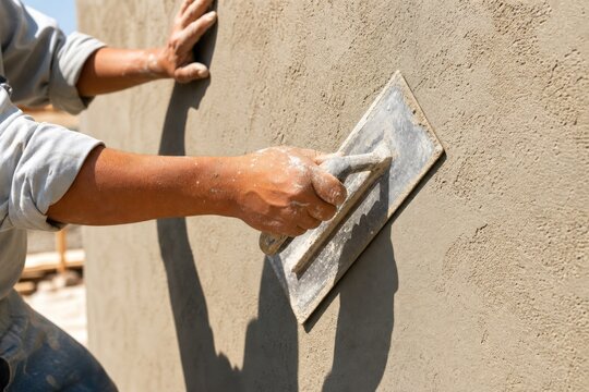 Professional builder applying wet plaster to concrete wall with metal trowel at outdoor construction site, close-up view of hands working diligently in bright sunlight.