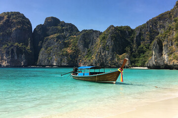 Obraz premium Thai longtail boat floats on turquoise sea beside white sandy beach, towering limestone cliffs under clear blue sky.