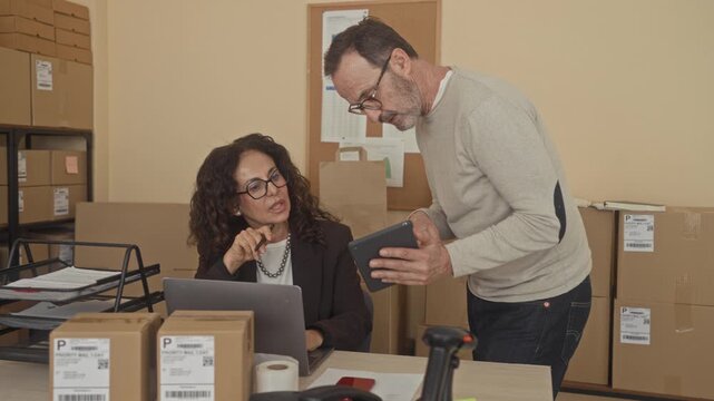 Man leans over showing a tablet to a seated woman at a packing desk with laptop, parcels, labels and scanner inside a building; concentration efficiency.
