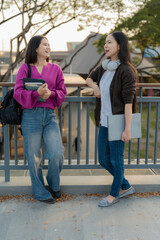 Fototapeta premium Two happy Asian student girls walking together on university campus, holding laptop and books while talking and gesturing.