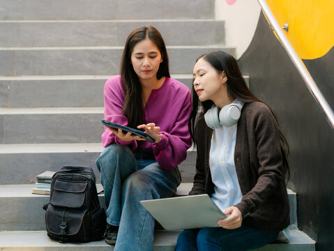 Two happy Asian student girls sitting on stairs, talking and studying together with laptop and tablet on university campus.