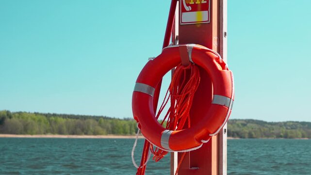 Bright red lifebuoy hangs on marina post with 112 emergency sign, rope, and metal fittings, set against calm blue water, distant shoreline, and clear summer sky