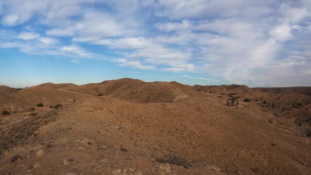 PAN: Expansive desert landscape near Matmata, Tunisia, with rolling arid hills, scattered shrubs, and dramatic cloud shadows creating contrast across the rugged terrain beneath a vast sky