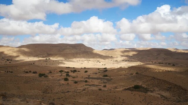 PAN: Expansive desert landscape near Matmata, Tunisia, with rolling arid hills, scattered shrubs, and dramatic cloud shadows creating contrast across the rugged terrain beneath a vast sky