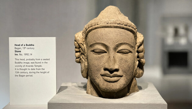 Ancient stone head of a buddha sculpture from the 13th century bagan period displayed on a white pedestal with an informational museum sign