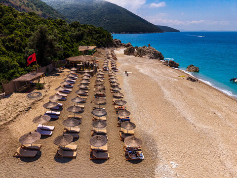 Aerial view of a beach with rows of straw umbrellas and sun loungers, a red flag flying, turquoise sea, and lush green hills in the background in Sarande, Vlore County, Albania.