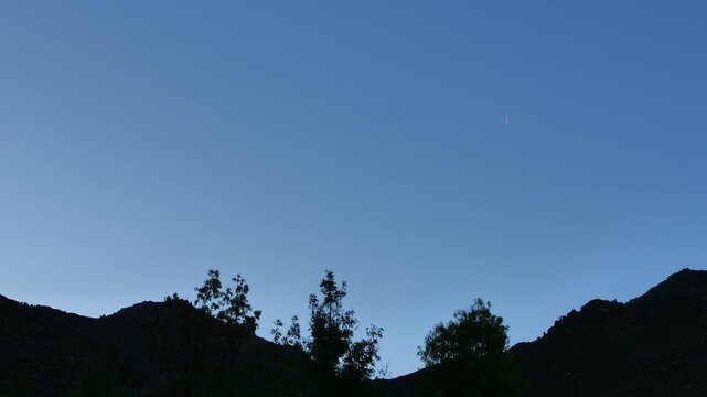 Small half moon on a pale blue sky with a mountain ridge in the foreground, 4K Time Lapse