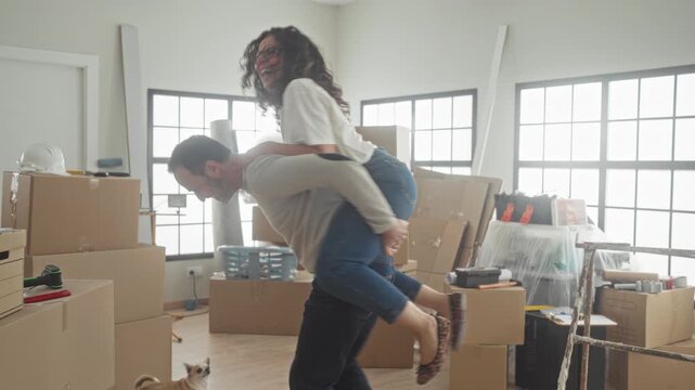 Man gives woman a piggyback, hands on shoulders and laughing amid stacked moving boxes, ladder and small dog in a bright building; joy togetherness.