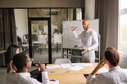 A facilitator leads an investment education session near whiteboard, discussing strategies with participants around a conference table in a contemporary office environment