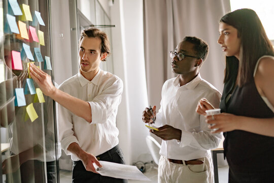 Diverse team of three, two man and woman actively engages in collaborative brainstorming session, sharing insights while utilizing colorful sticky notes on a glass board in a bright, modern workspace