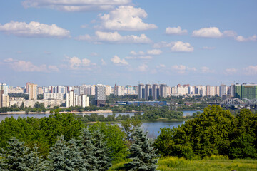 Fototapeta premium Kyiv, Ukraine, a panoramic view across the Dnipro River, revealing the city's left bank residential areas, modern high-rise buildings, and the Darnytskyi railway bridge under a clear sky with clouds