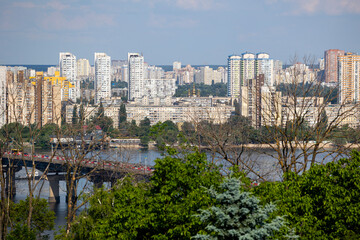 Fototapeta premium Kyiv, Ukraine, a panoramic view of the Paton Bridge spanning the Dnipro River, with bustling residential districts and modern high-rise buildings lining the scenic left bank under a clear sky