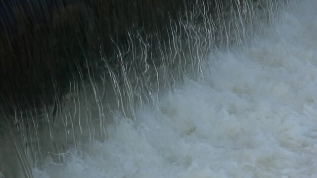 A steady, continuous stream of thick white froth and river water flowing downstream from a rubber weir in relaxing slow motion.

