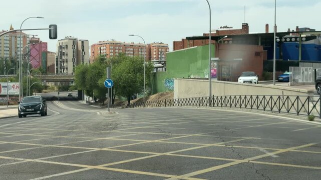 MADRID, SPAIN, June 24-2026:Urban street view with traffic signals and road signs. Concrete barriers line the road. Buildings rise in the distance. Green trees grow along sidewalks. Blue arrow sign di