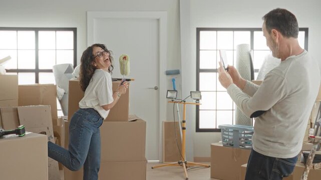 Woman holding paint roller and smiling while man photographs her with smartphone amid packed moving boxes and tools inside building; playful.