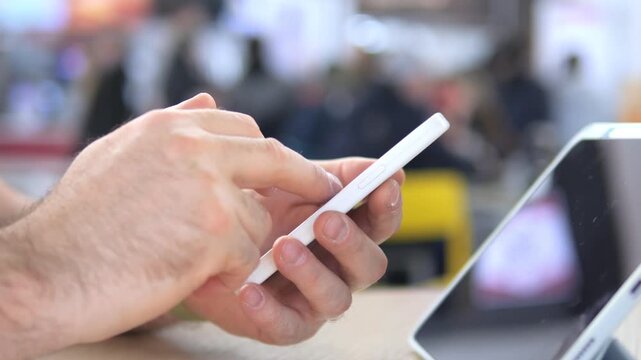 Hand holds white smartphone, fingers tap screen. Blurred background suggests public space. Person focused on device, intent and calm. Ideal for tech, communication, or digital lifestyle themes