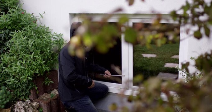 A person in dark clothing uses a tool to pry open a white window on a white house exterior wall
