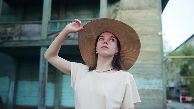 Young woman adjusting straw hat outside a weathered wooden building in soft daylight