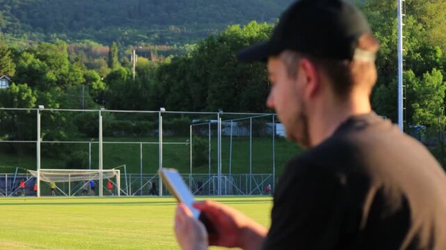 White man using smartphone by football field at sunset with athletes training in the background. Concept of phone addiction, digital distraction, social isolation, procrastination, modern screen