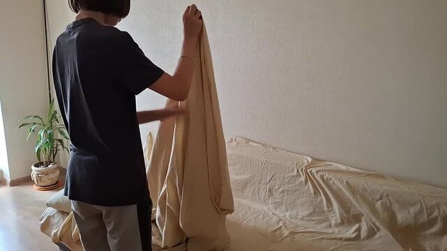 A teenage boy straightens the sheets and blankets on a bed in a simple room